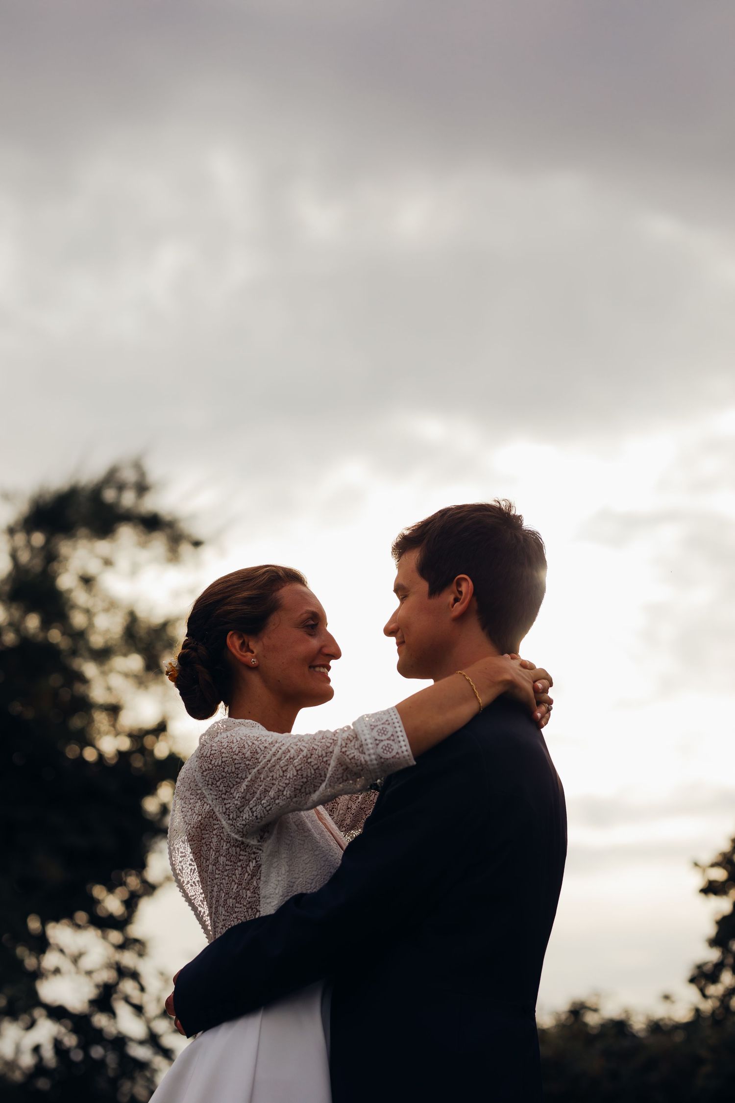 Bride and groom embracing romantically at dusk with dramatic cloudy sky and trees behind them.