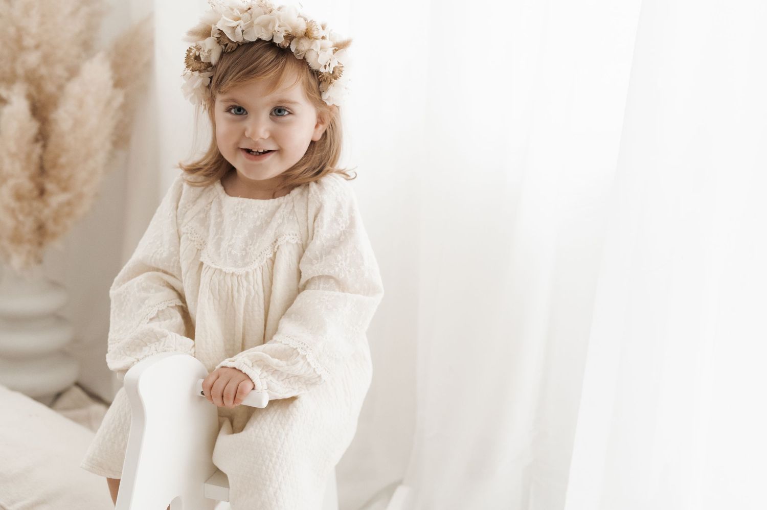 Child in vintage white dress and floral crown sitting against light backdrop.