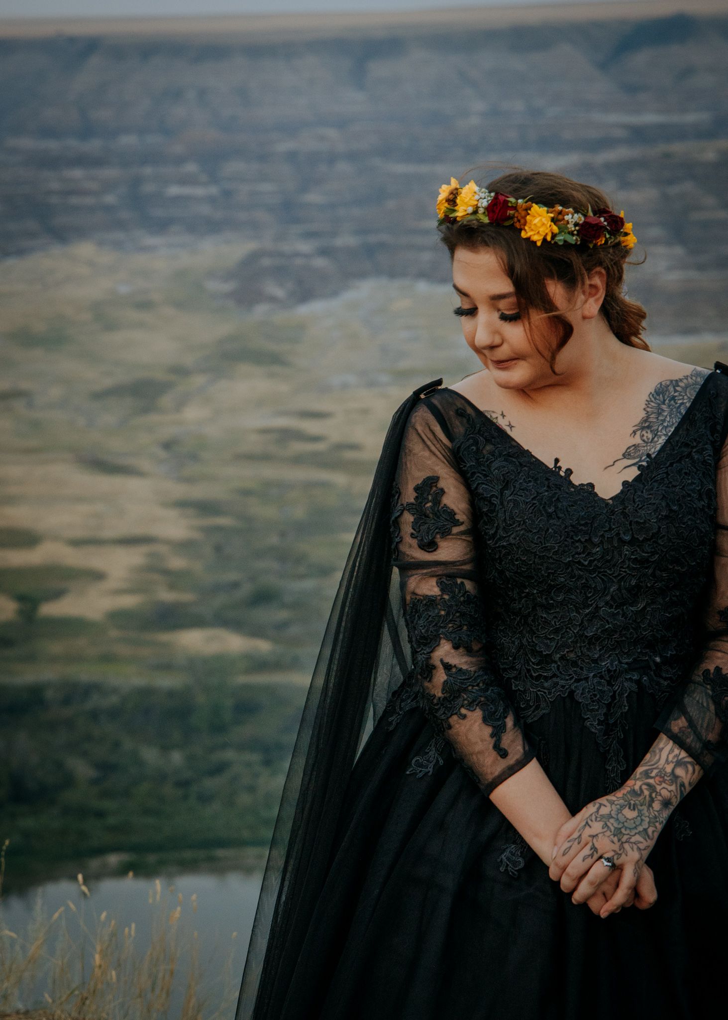 A person wearing a black lace dress with a floral crown stands against a scenic mountain landscape backdrop.