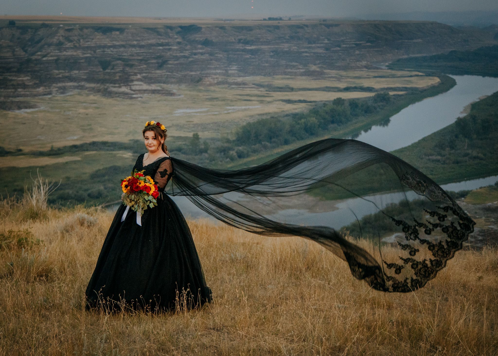 A figure in a dramatic black gown with flowing cape stands on a cliff overlooking a winding river valley.