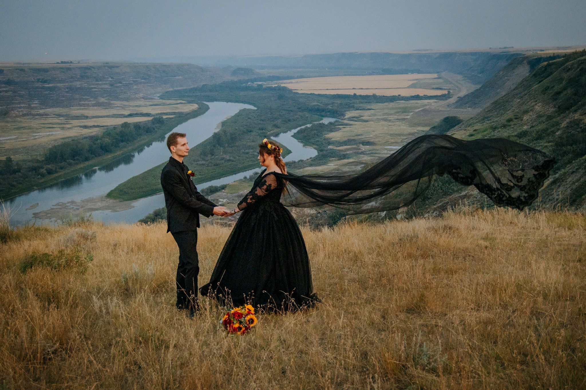 A couple in formal attire stands on a hilltop overlooking a scenic river valley during golden hour.