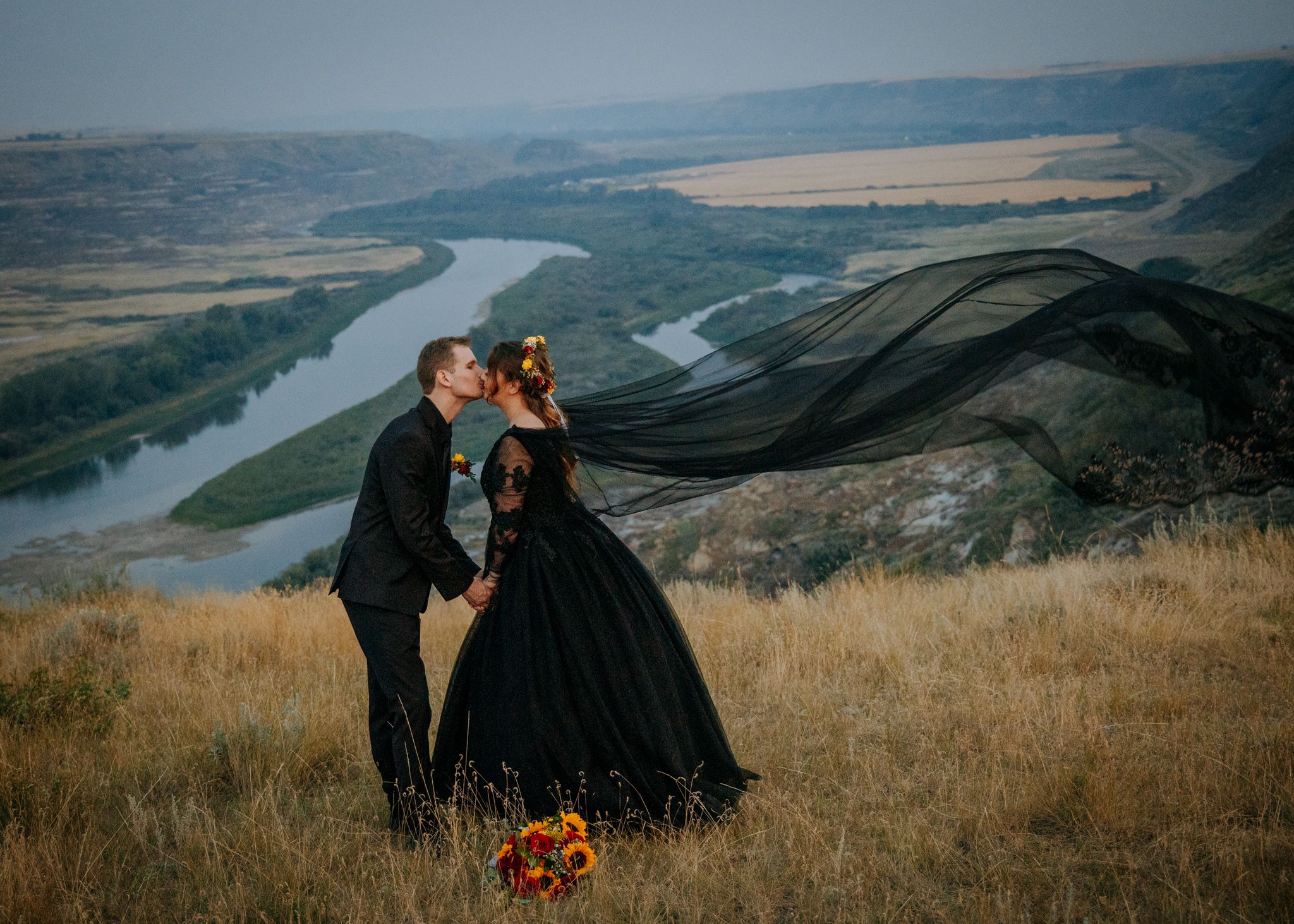 A dramatic black dress flows in the wind on a hilltop overlooking a winding river valley below.