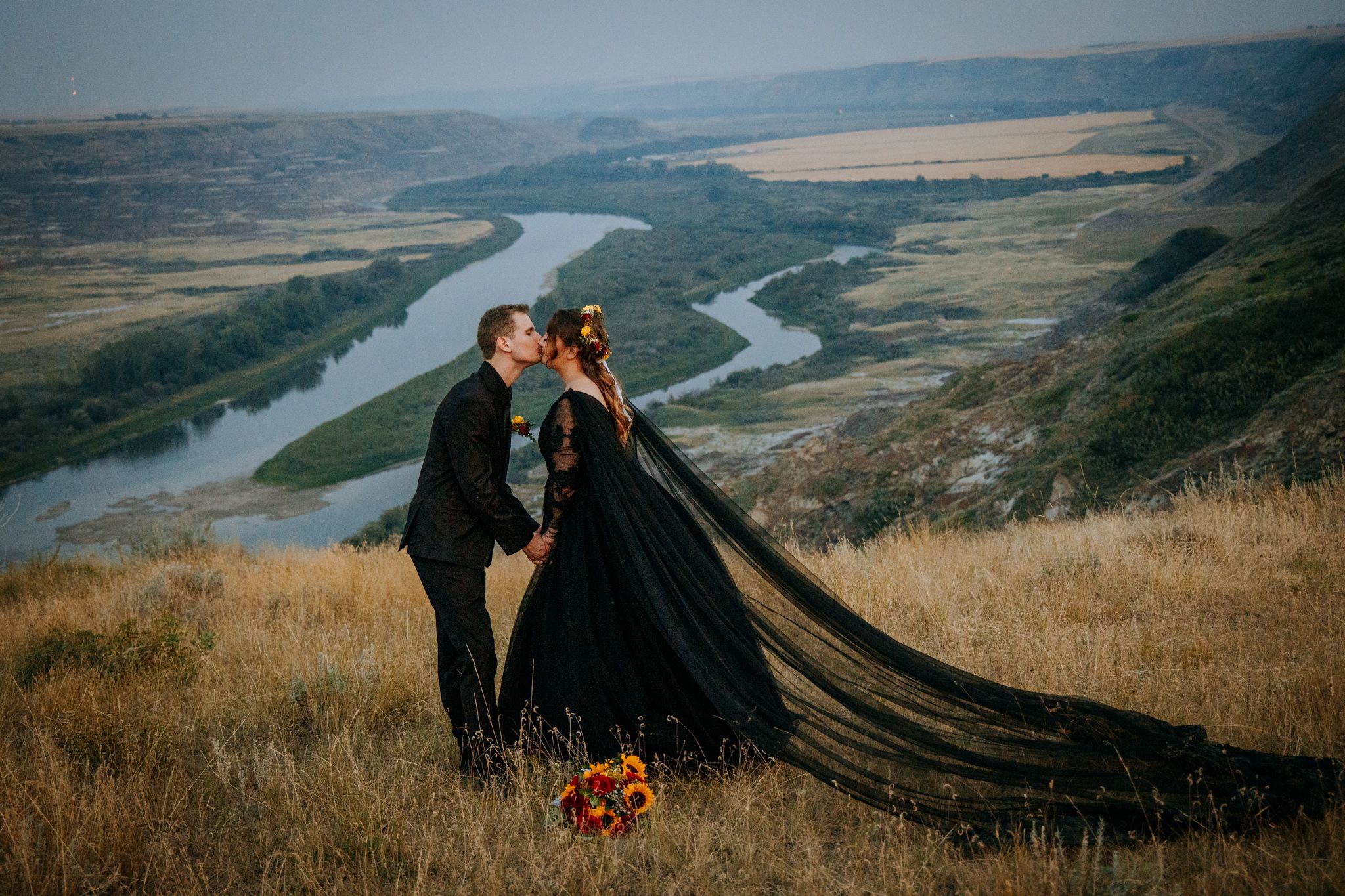 A couple shares an intimate moment overlooking a scenic river valley with winding roads and rolling hills in the background.