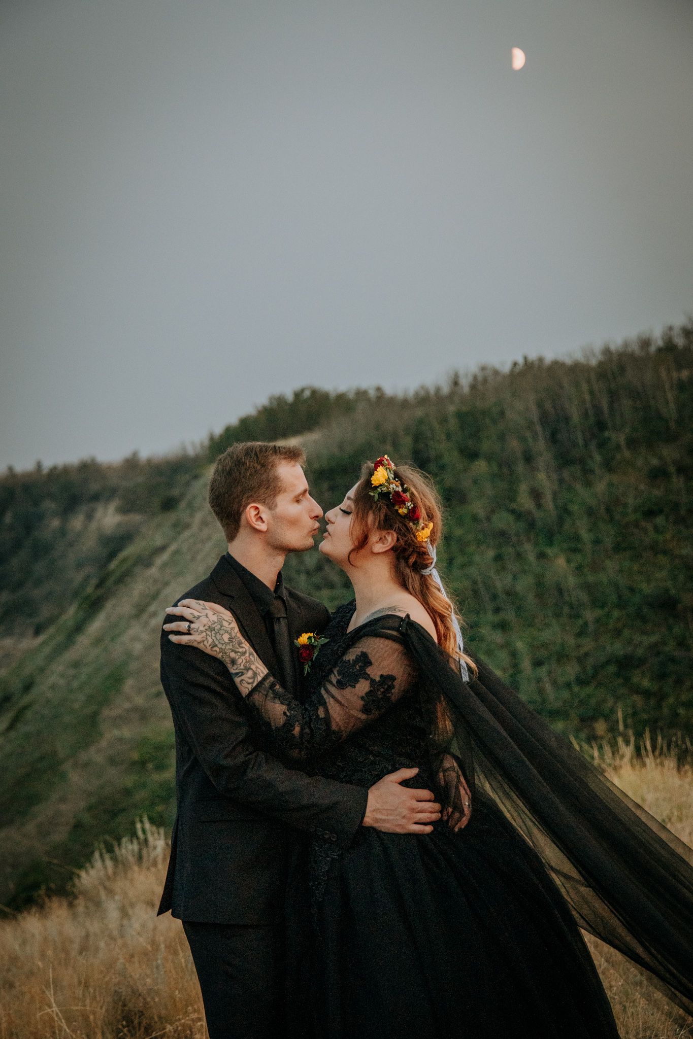 A romantic couple in black gothic wedding attire embrace on a hillside at dusk with the moon visible in the sky.