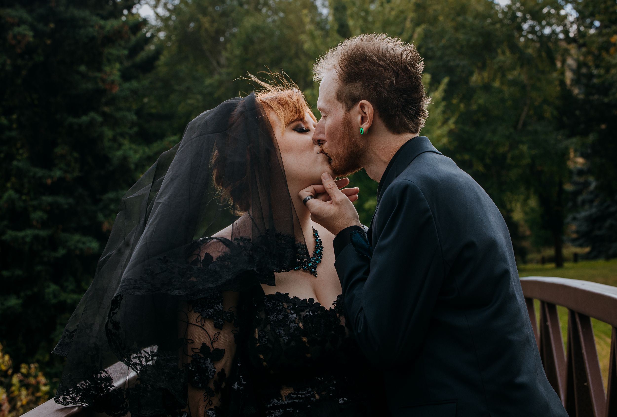 A couple in dark attire share an intimate moment on a bridge surrounded by trees.