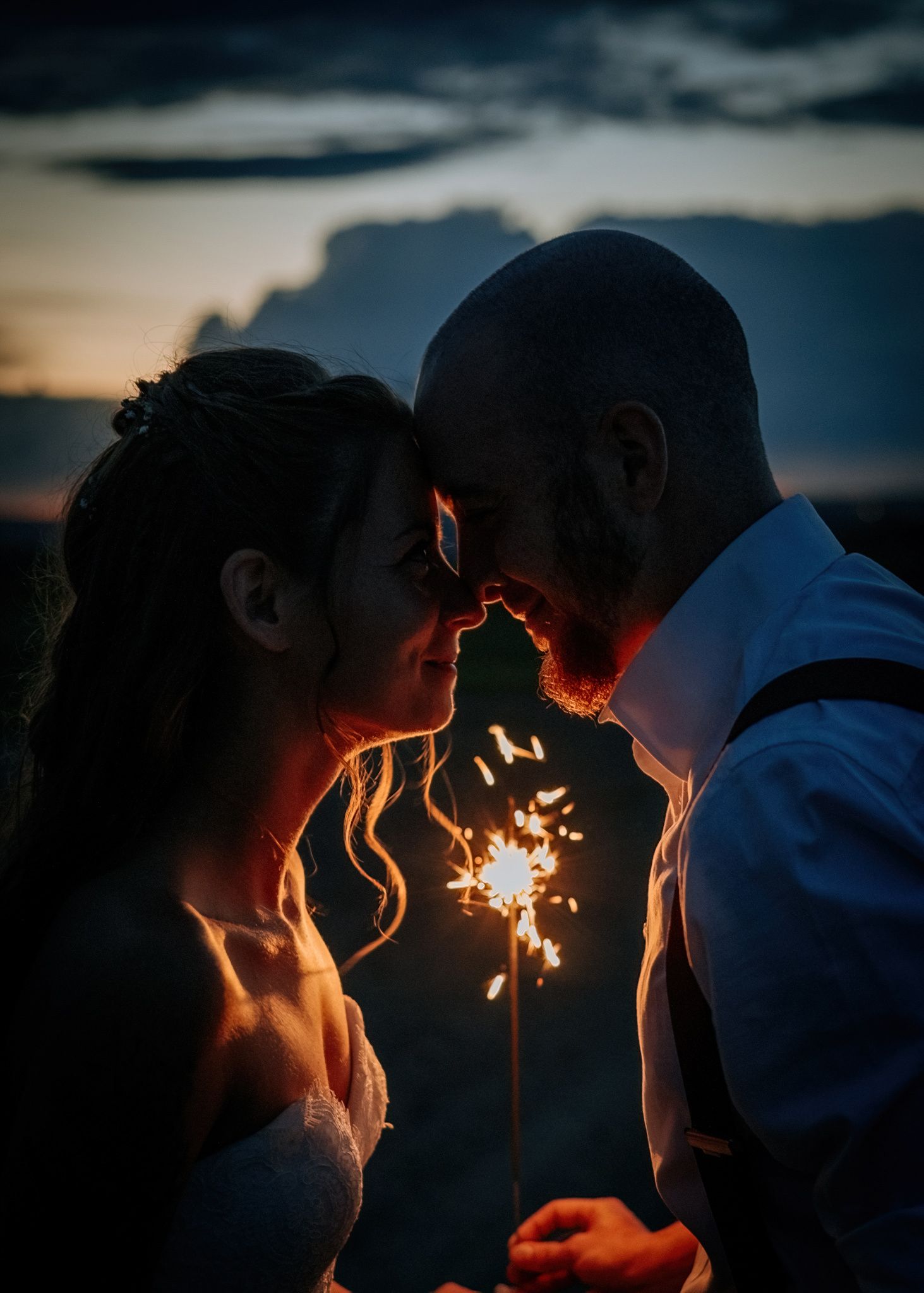 A couple shares an intimate moment lighting sparklers together at dusk against a moody sky backdrop.