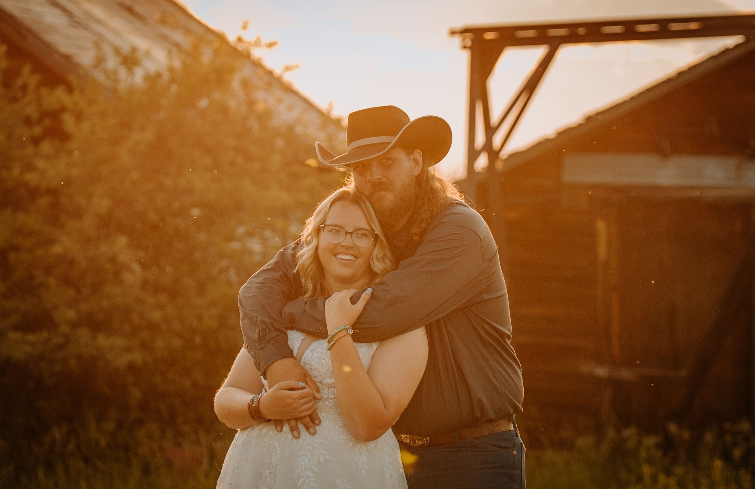 A couple embraces in a warm golden sunset glow with a rustic wooden structure in the background.