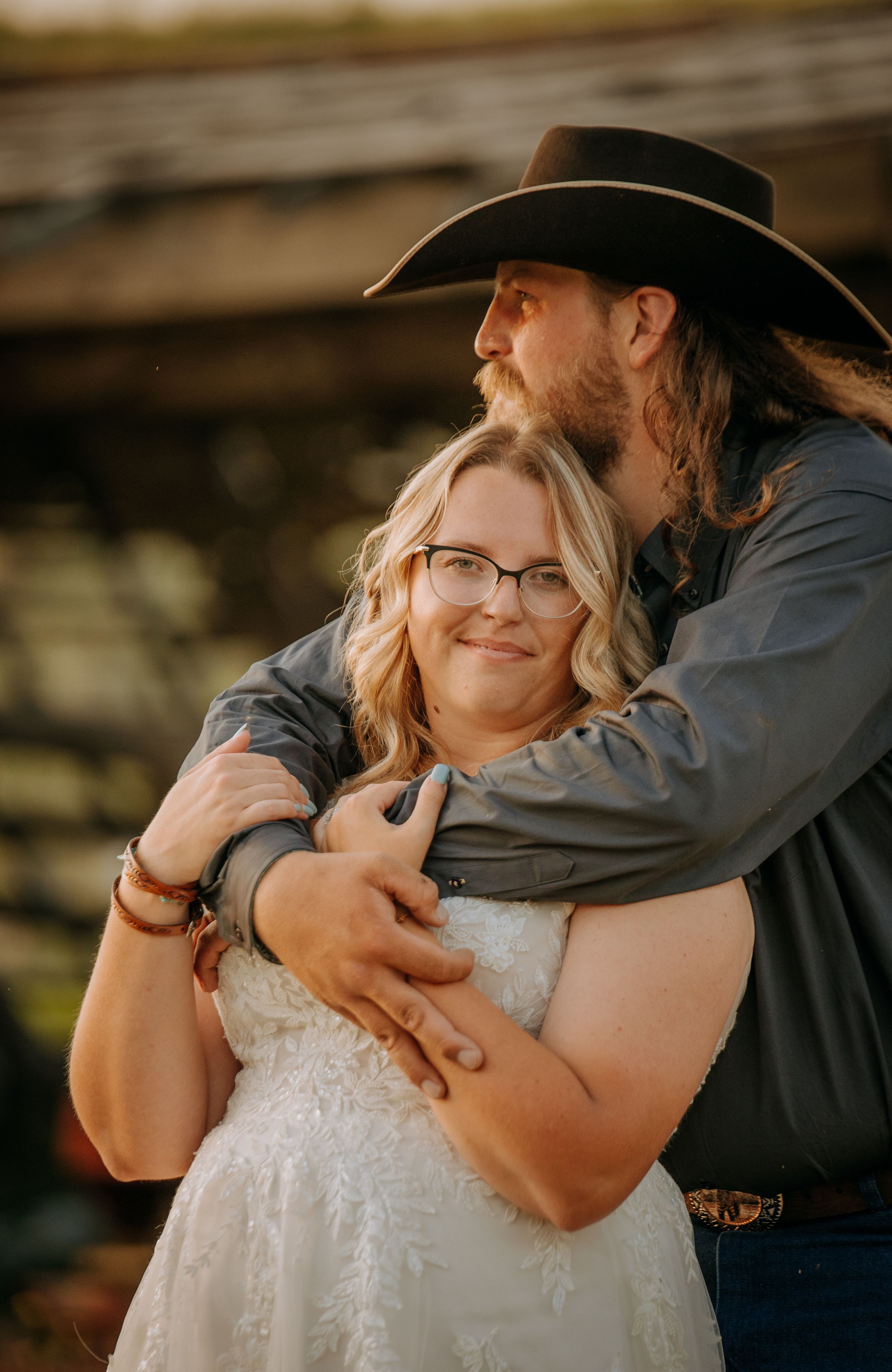 A romantic couple embraces outdoors at sunset, with the person in a black hat holding their partner in a white lace dress.