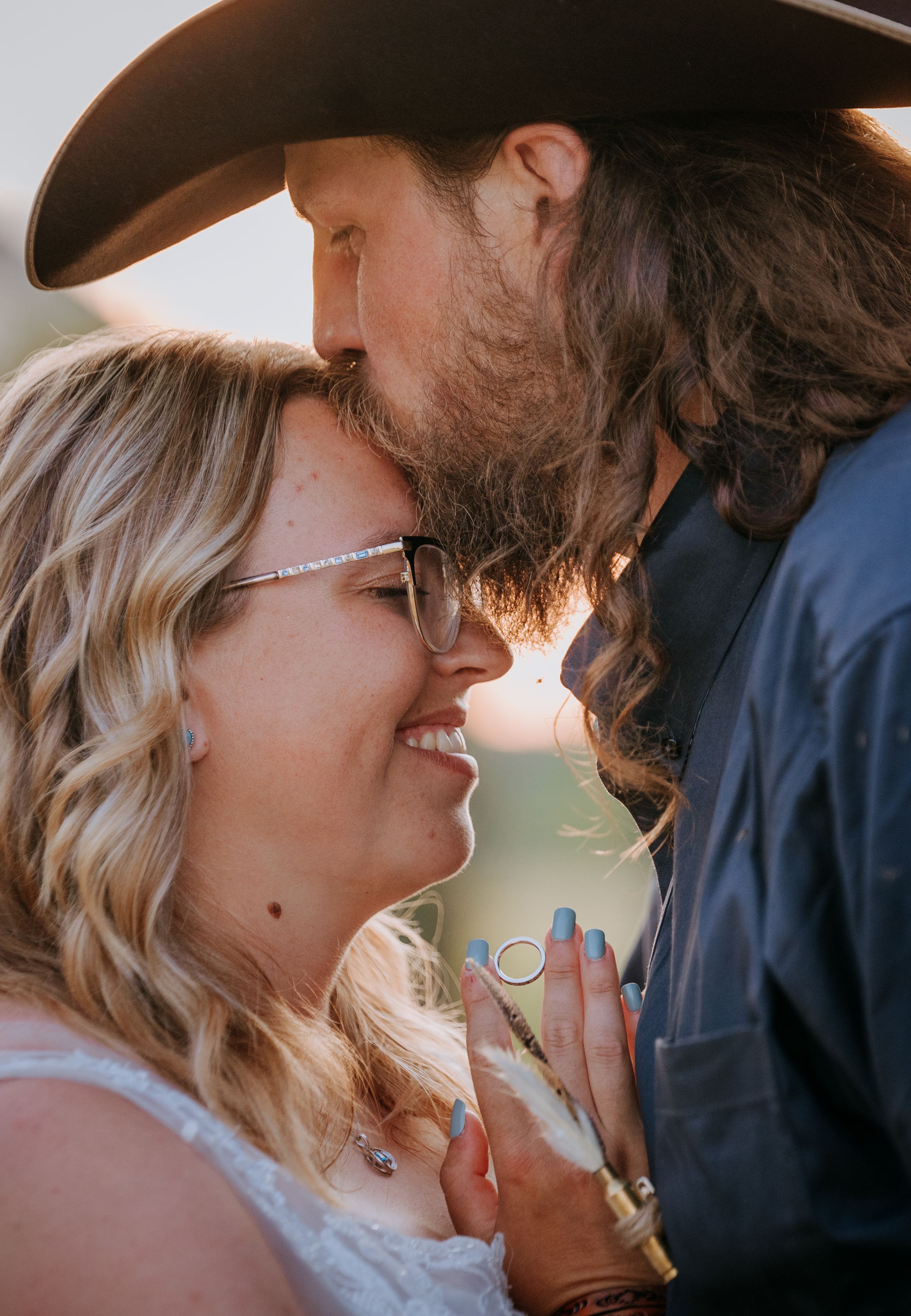 A romantic moment captured at sunset between a bearded man in a cowboy hat and a woman in glasses smiling together.