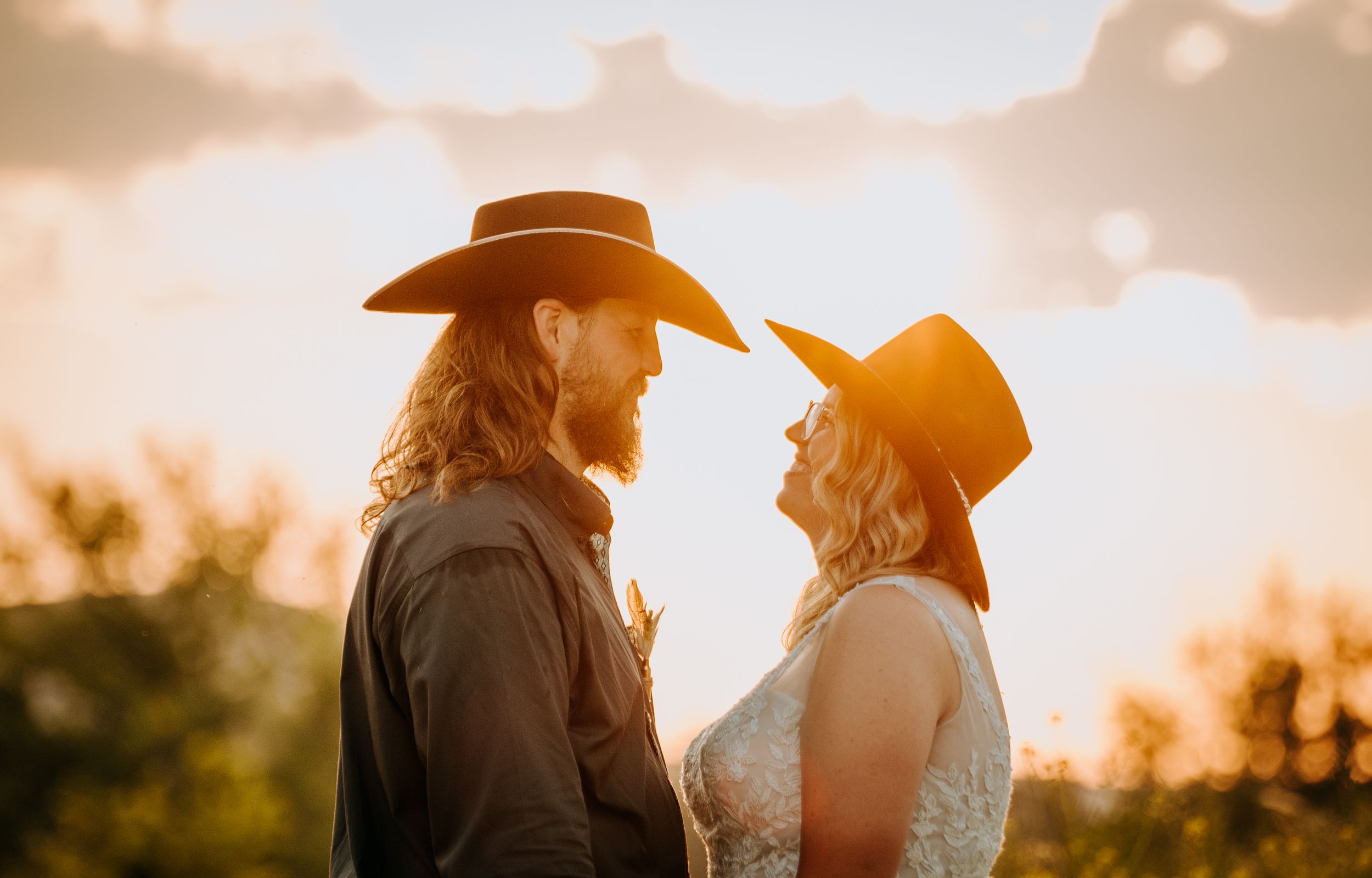 Silhouetted couple in cowboy hats share tender moment at sunset with golden light streaming through trees.