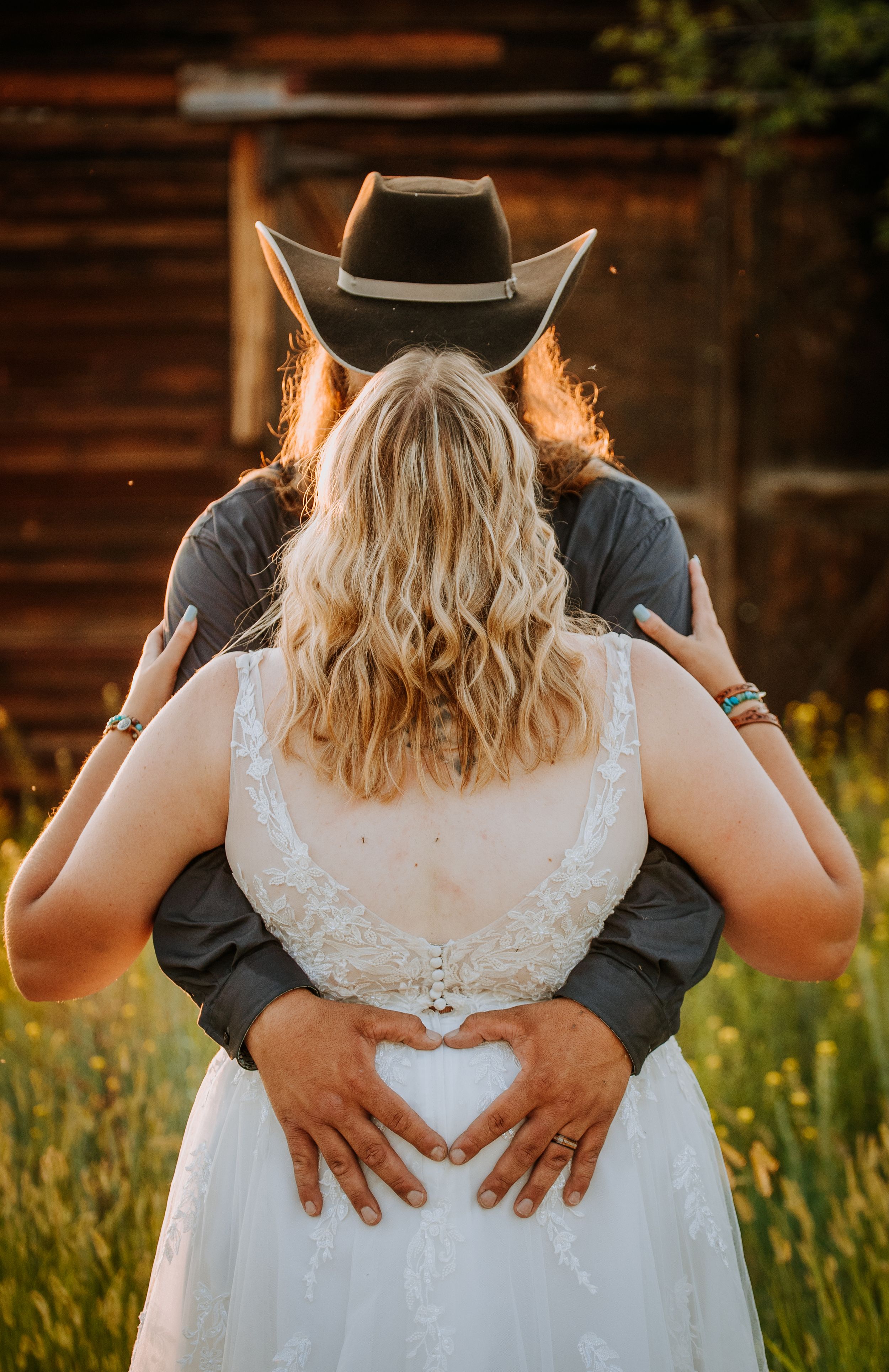A couple in wedding attire embraces in a golden field at sunset, with hands forming a heart shape on the bride's back.