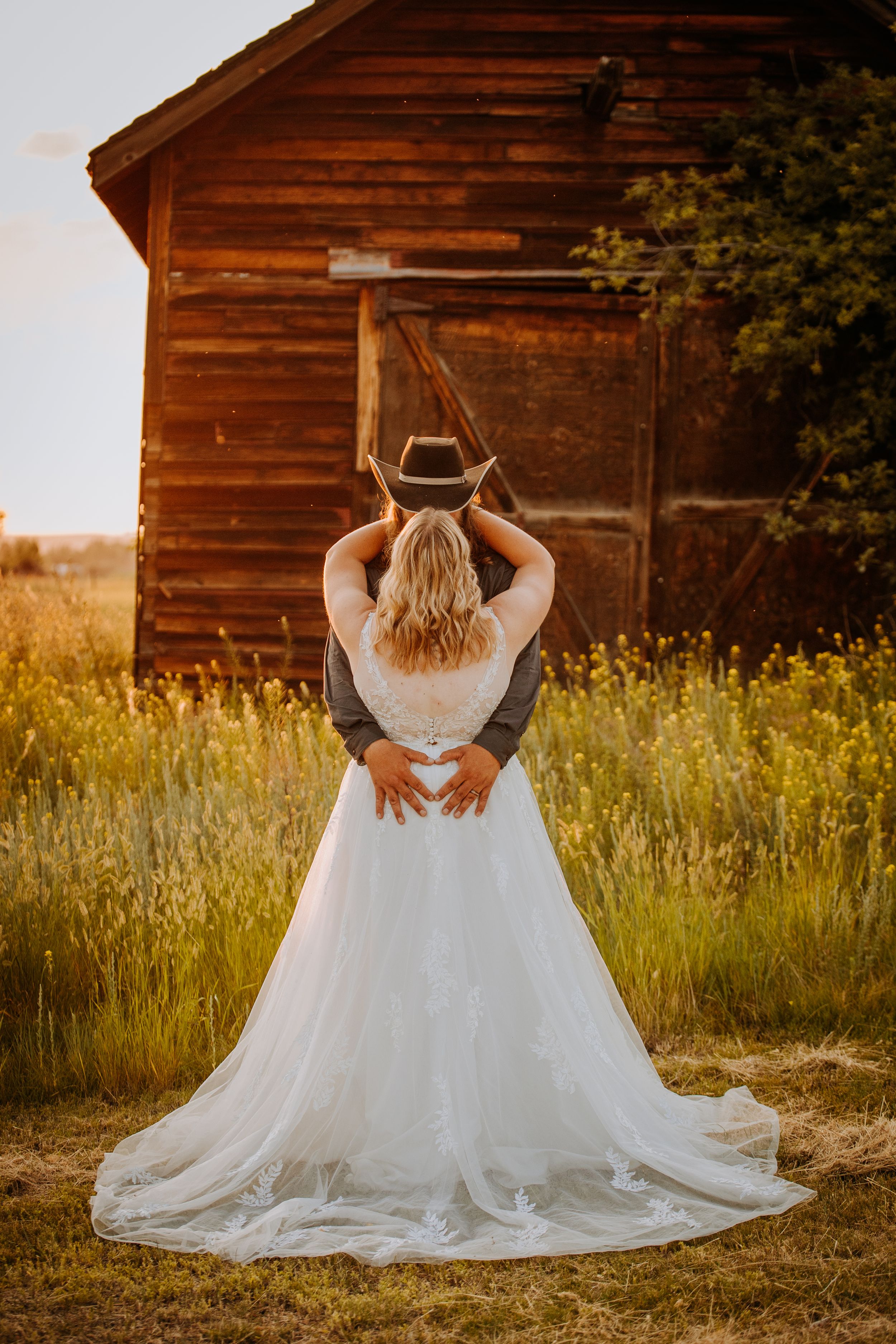 A person in a flowing white wedding dress sits in a golden meadow at sunset beside a rustic wooden barn.