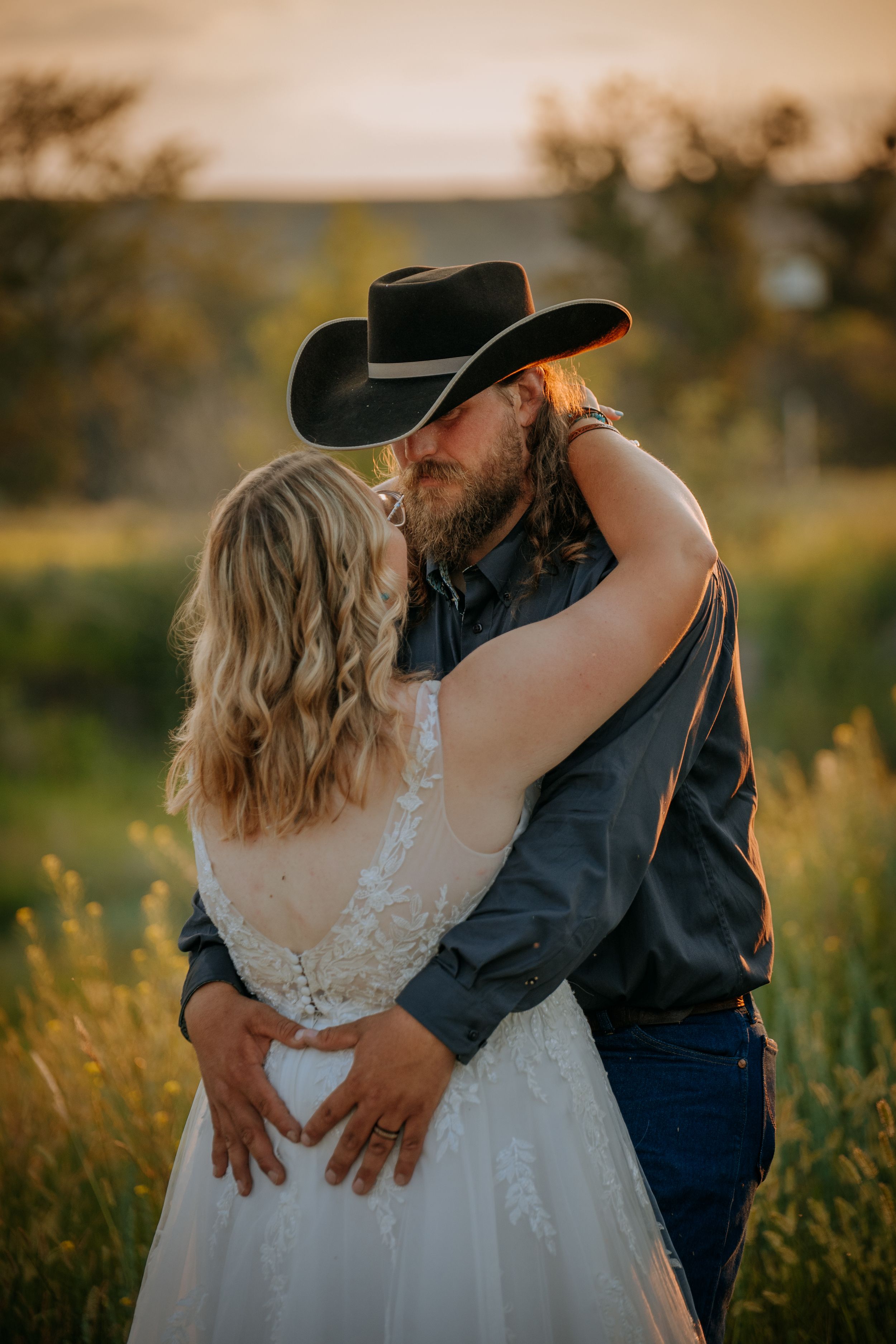 Romantic couple embraces in a golden field during sunset while wearing western attire and wedding dress.