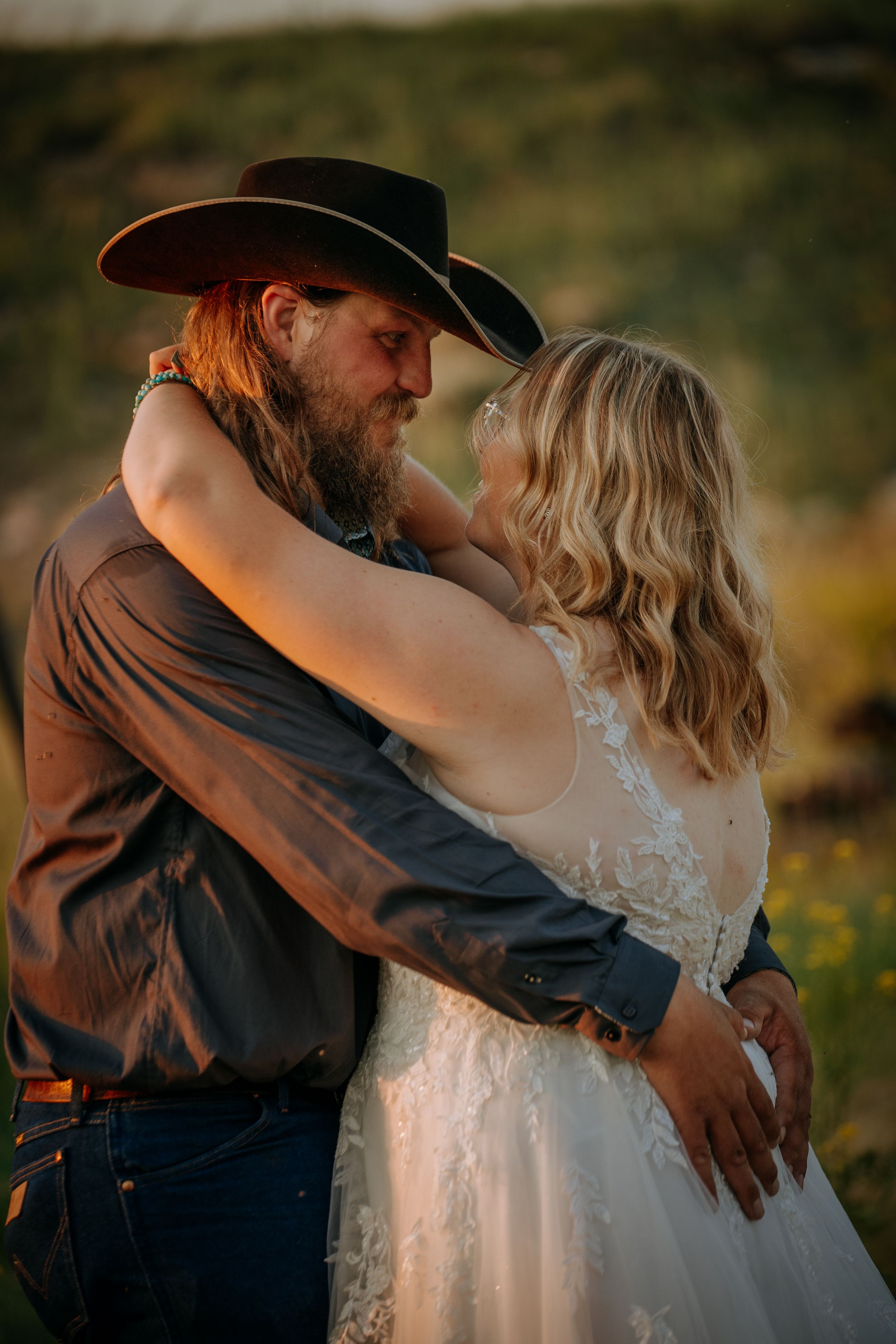 Romantic western-style wedding photo shows couple embracing at sunset with cowboy hat and lace dress.