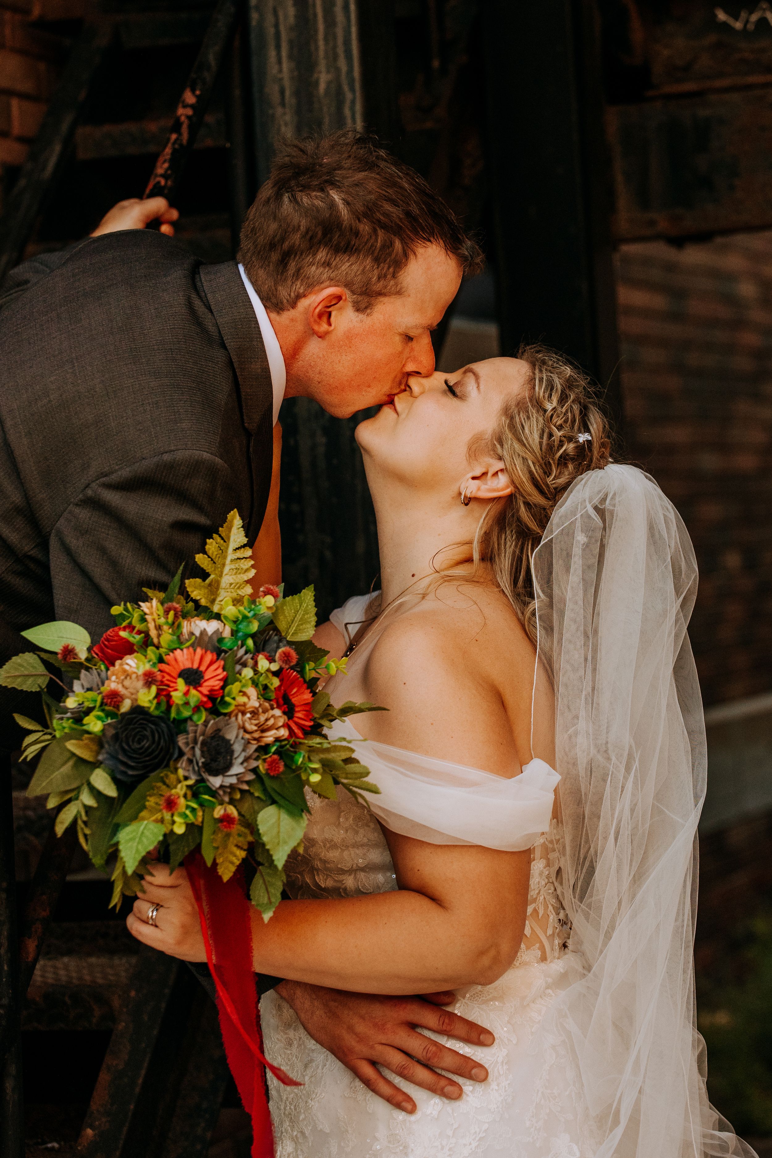 Romantic wedding couple shares intimate kisses in moody lighting with vibrant red floral bouquet accents.