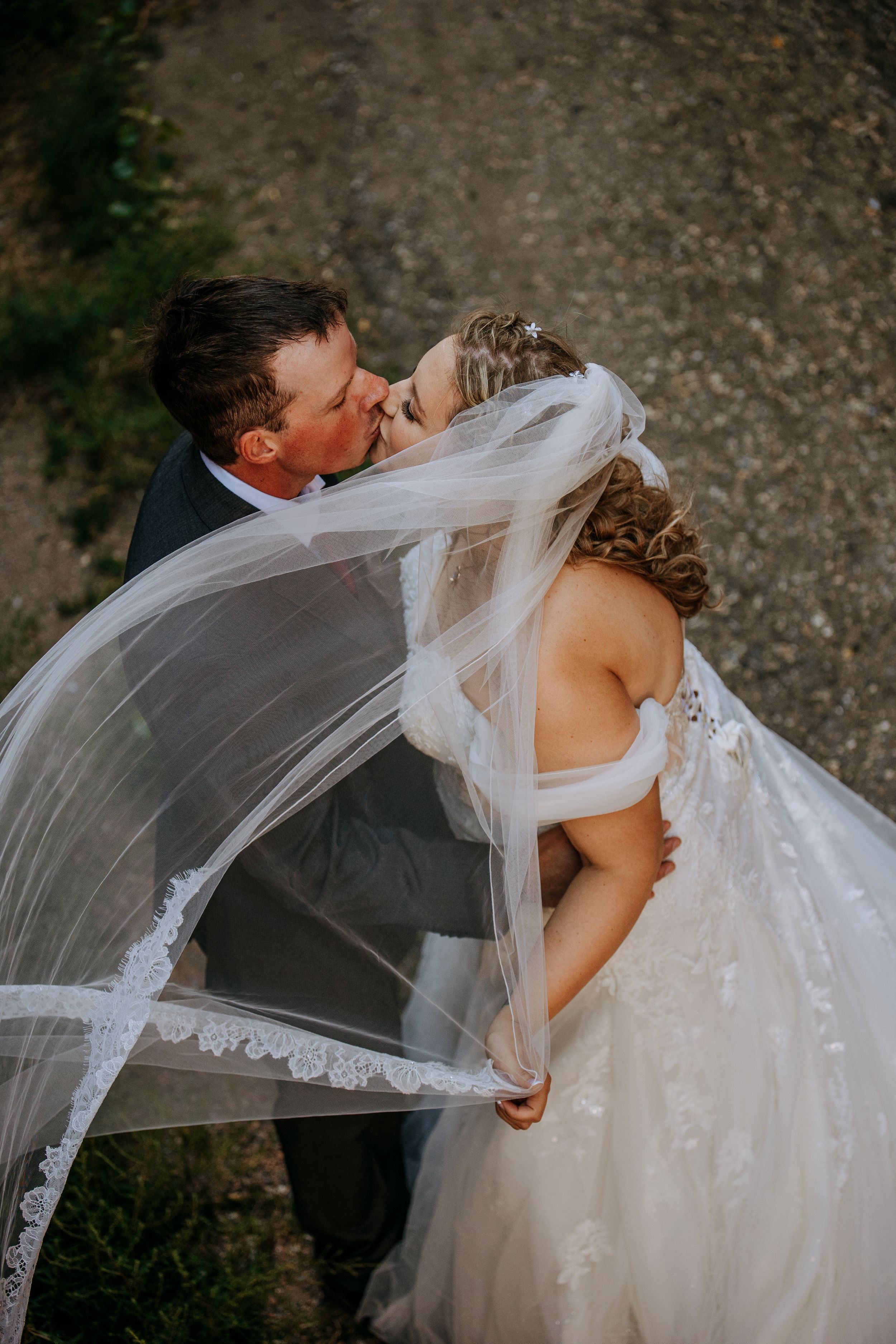 A romantic wedding couple embraces in a kiss as the bride's delicate lace veil flows dramatically in the breeze.