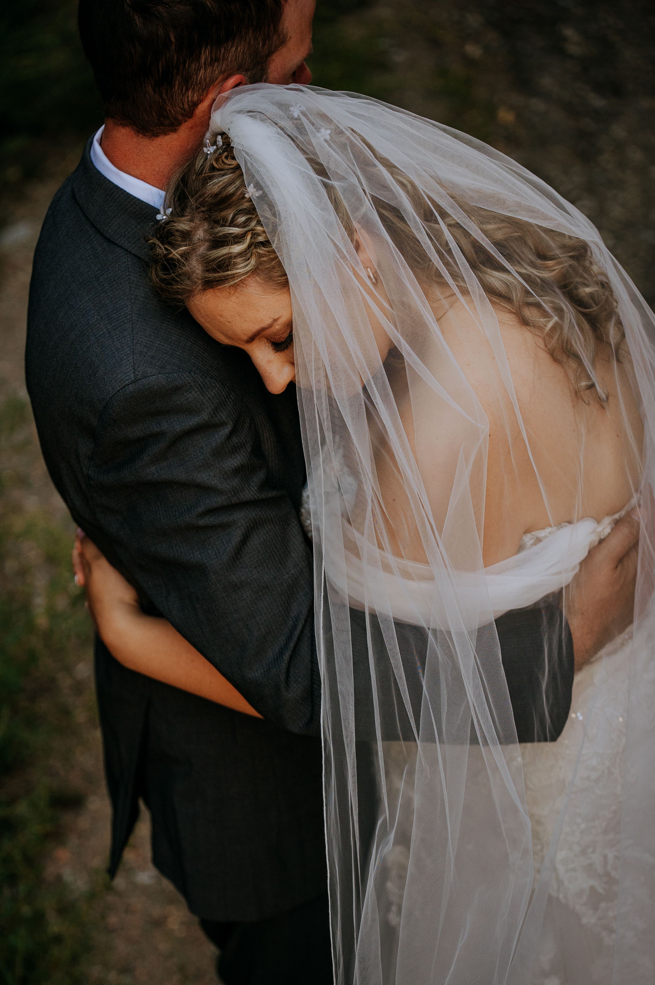A romantic wedding couple embraces outdoors as the bride's flowing veil catches the light in an intimate moment.