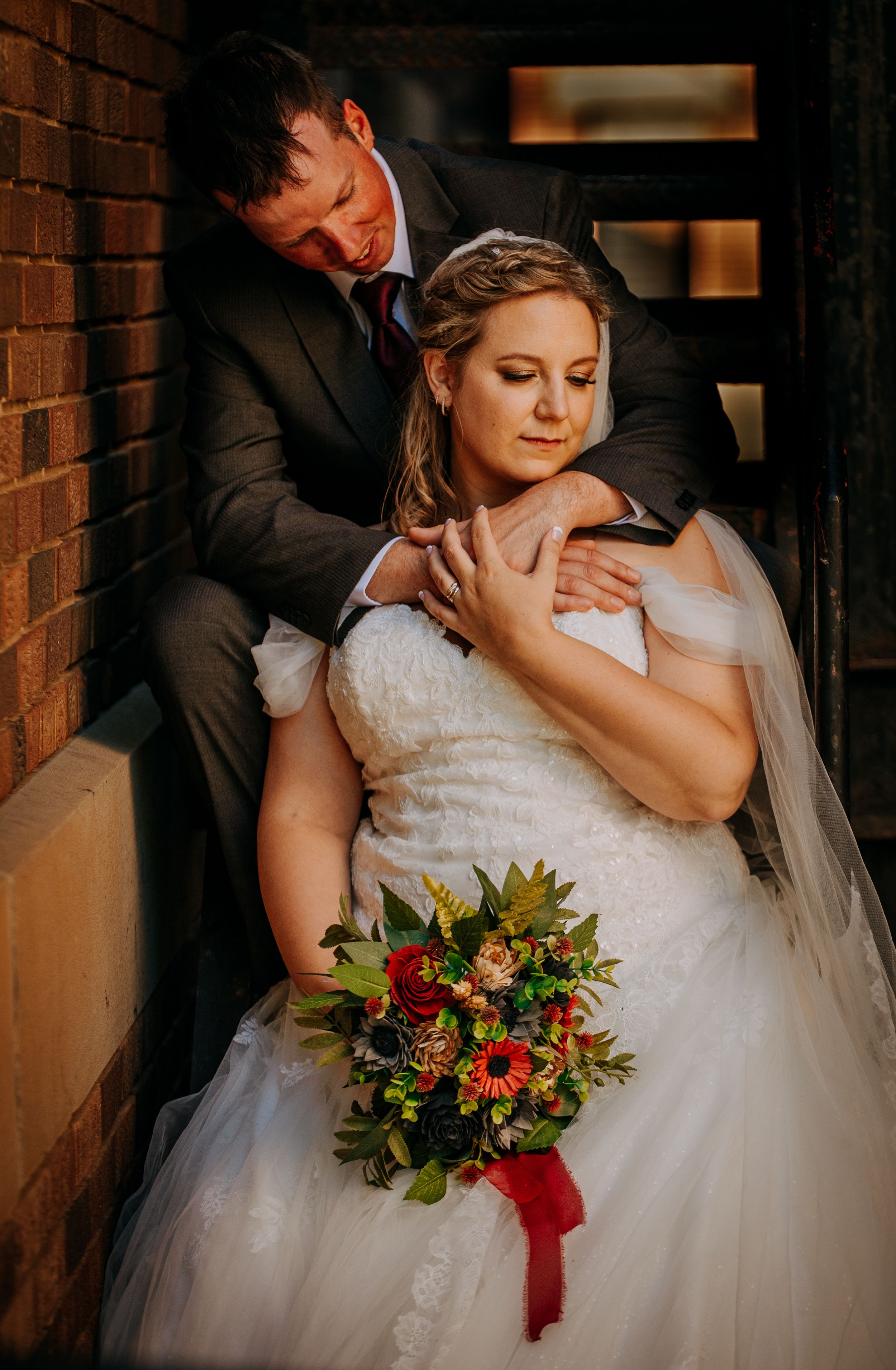 Romantic wedding couple embraces against brick wall backdrop with bride holding vibrant autumn bouquet.