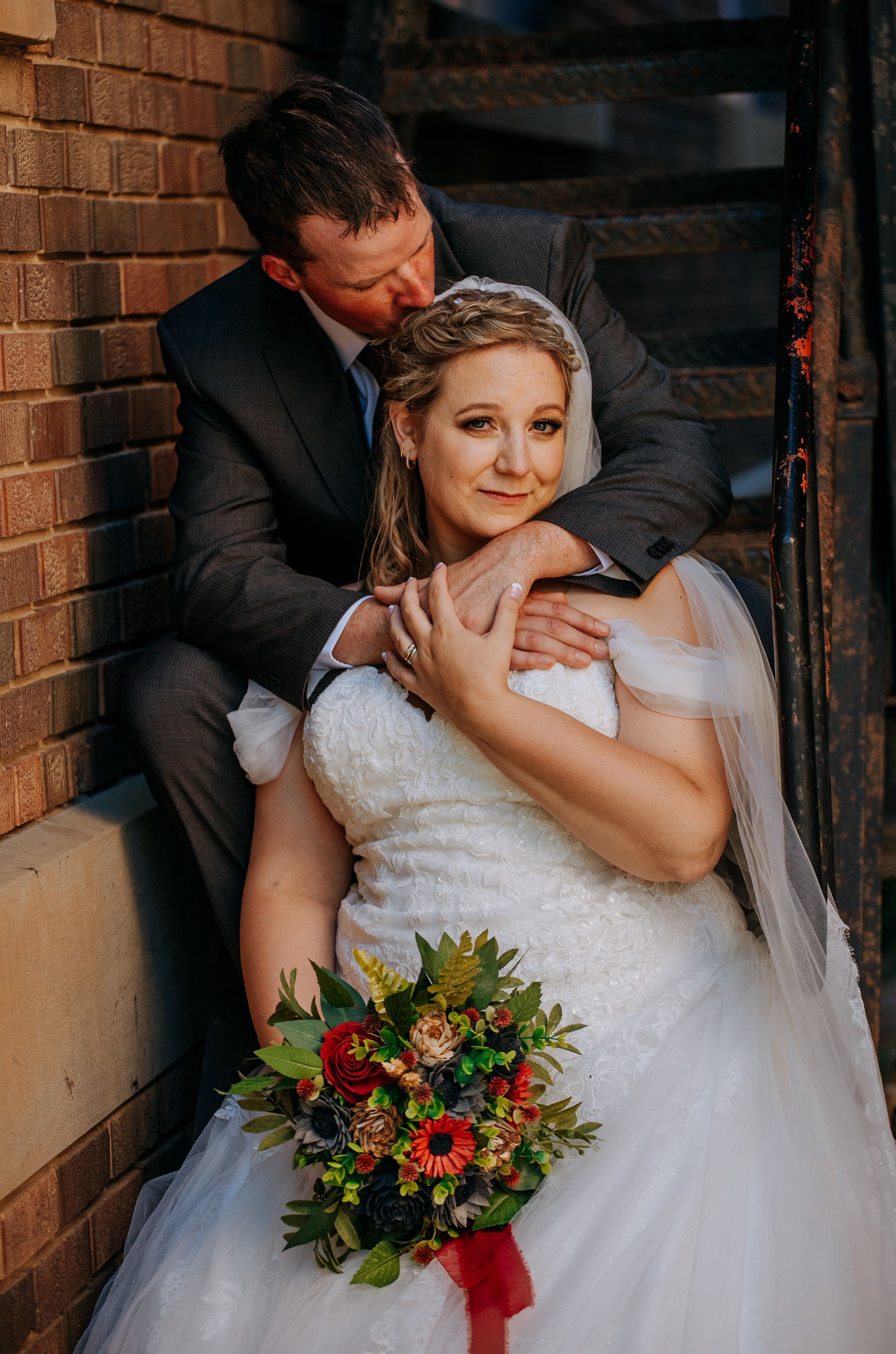 A romantic wedding couple embraces in front of a brick wall while holding a vibrant red and green bouquet.