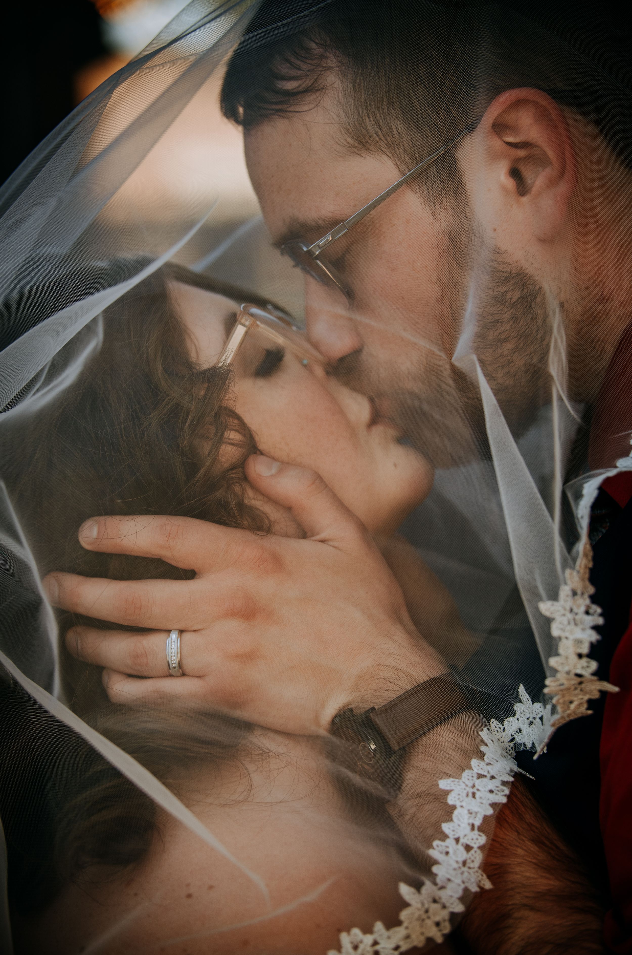 Romantic wedding couple shares intimate kiss under delicate lace veil in soft lighting.