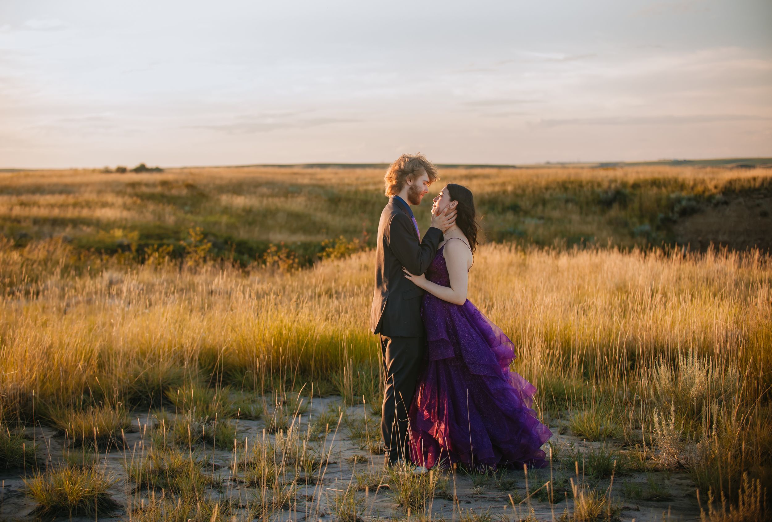 A couple sits together in a golden prairie field during sunset while wearing casual attire.