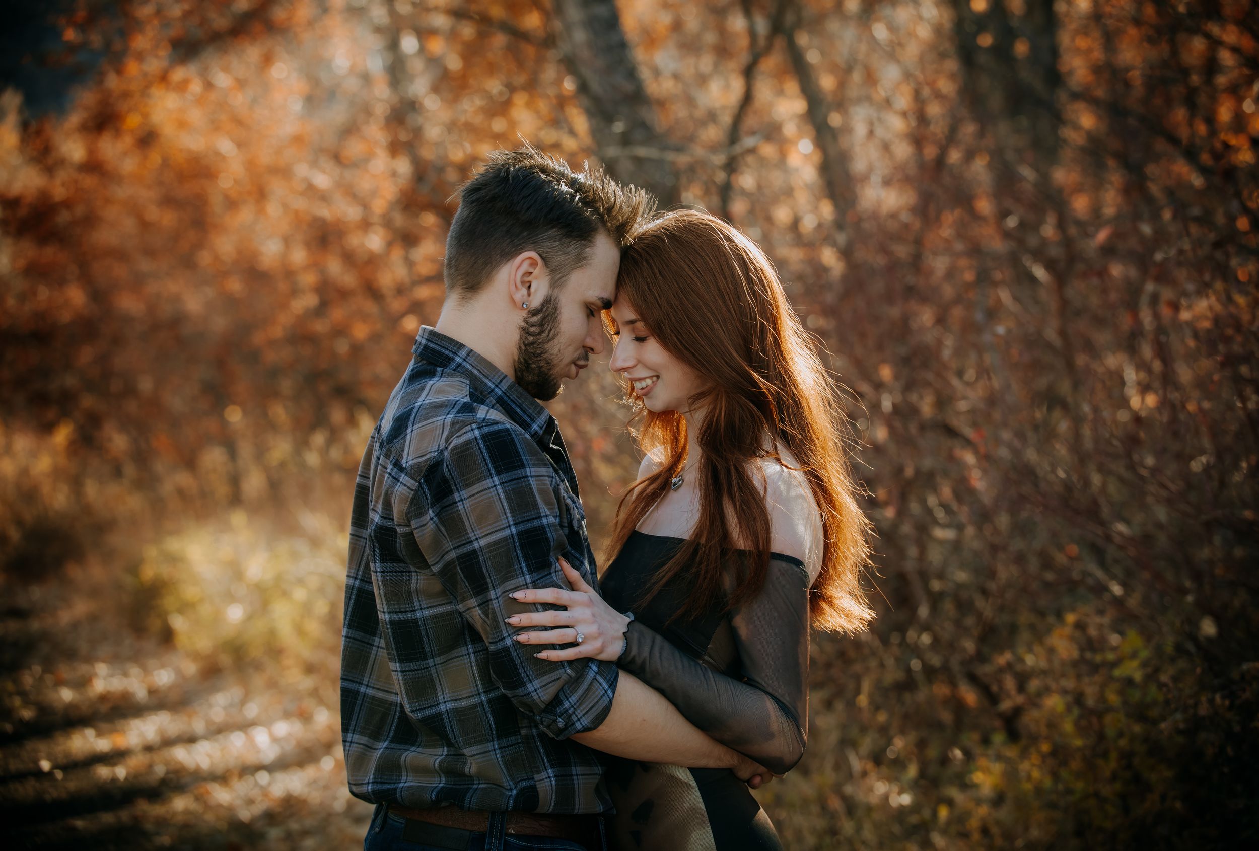 A couple embraces in a romantic autumn forest setting with warm golden sunlight filtering through the trees.