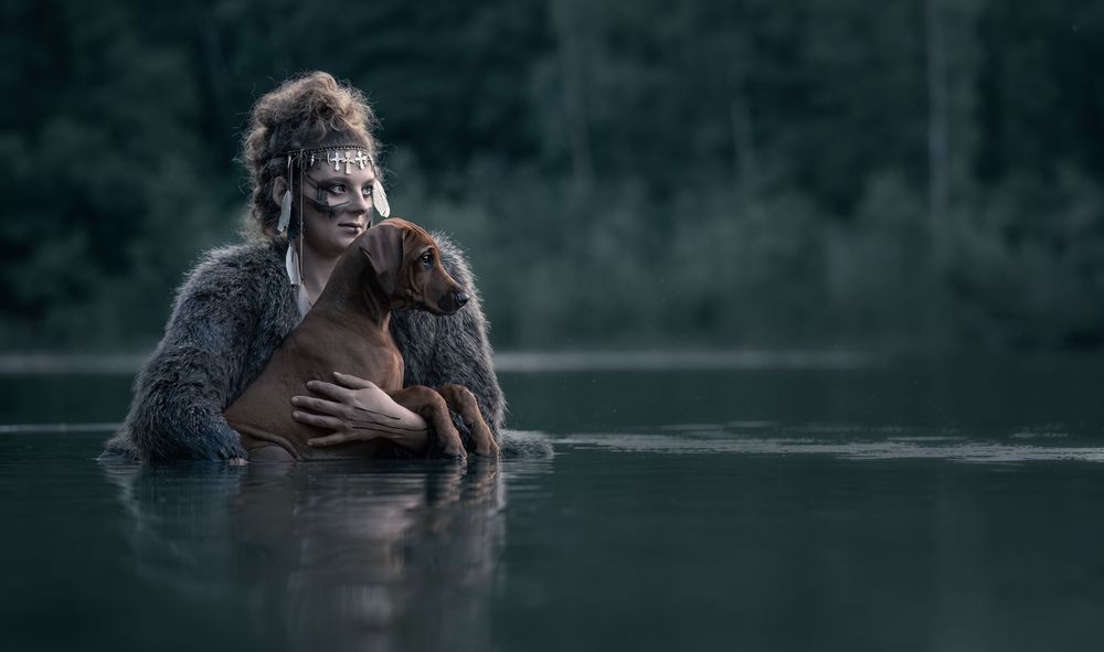A dramatic viking portrait showing a woman with a ridgeback dog in the water at dusk.