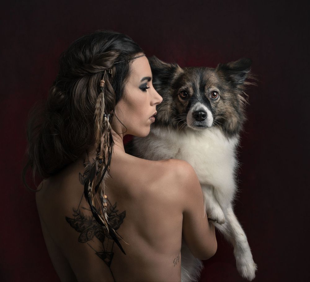Artistic portrait showing a person with long dark hair and a Shetland Sheepdog against a dark red background.