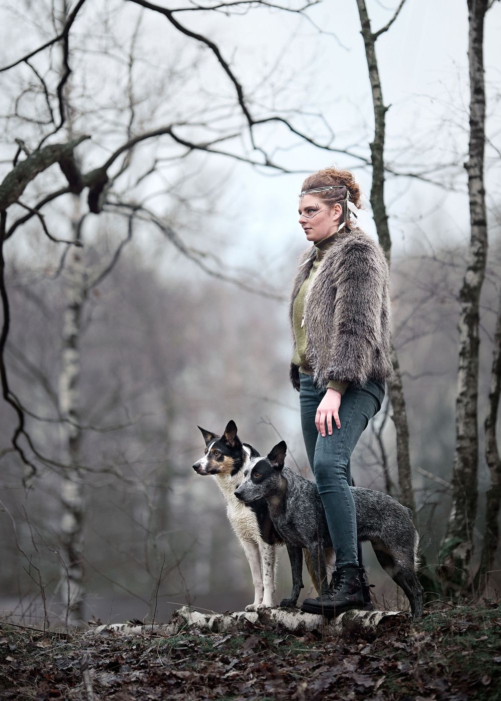 A person in a fur coat stands with two dogs in a misty winter forest setting.