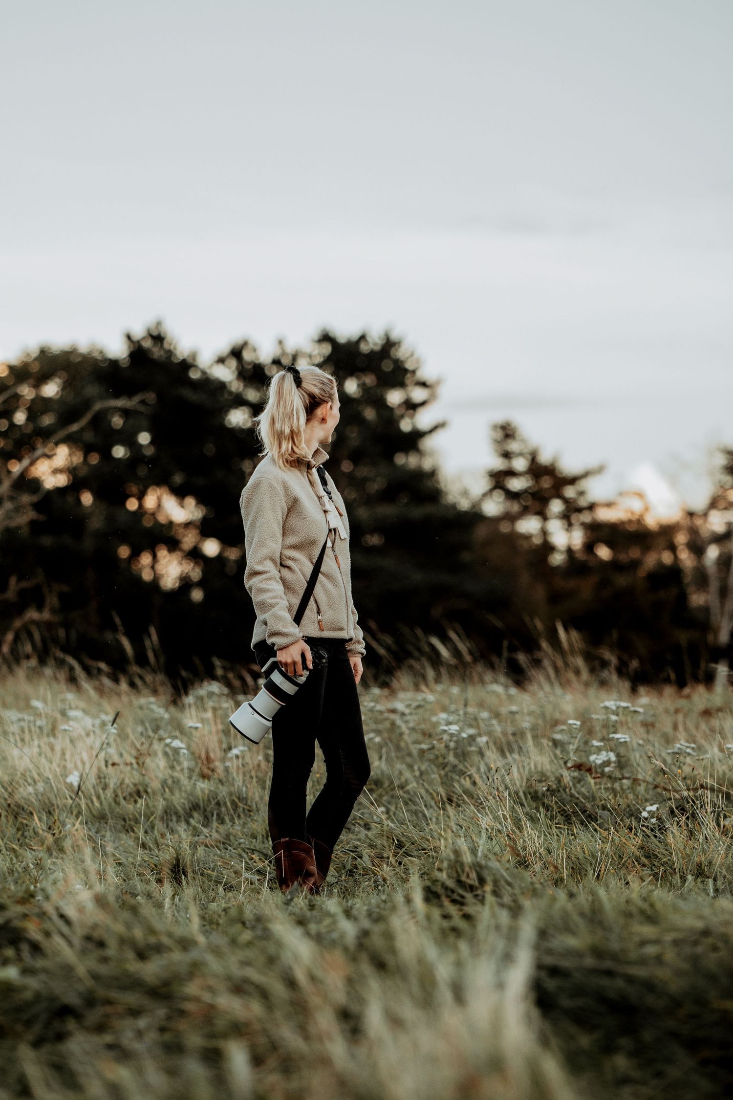 A photographer stands in a grassy field at sunset holding a camera, wearing a beige sweater and dark pants.