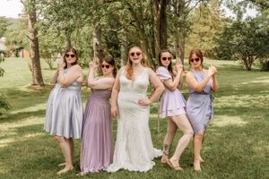 Bridesmaids in pastel purple and blue dresses pose together on a sunny lawn.
