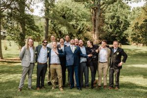 Groomsmen gather for a group photo in a mix of suits and casual wear on a green lawn.