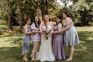 Bridal party wearing coordinated lavender and grey dresses stands together in a park setting.