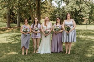 Wedding group photo with bridesmaids in pastel purple dresses on a grassy lawn.