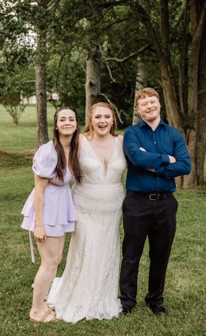 Wedding group photo showing bride in lace gown with guests in casual attire in outdoor garden setting.