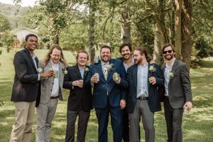 Three groomsmen in formal attire pose playfully for photos in a wooded outdoor setting.