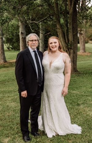 Outdoor wedding portrait with bride in lace dress and guests in formal black attire among trees.