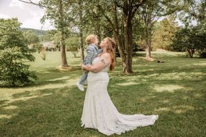 Bride in flowing white dress lifts child in joyful moment outdoors.