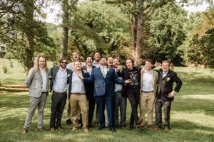 A group of groomsmen in casual and formal attire pose together on a sunny lawn with trees in the background.
