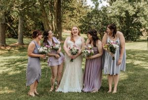 Wedding party in purple and gray dresses poses together on a grassy lawn with trees in the background.