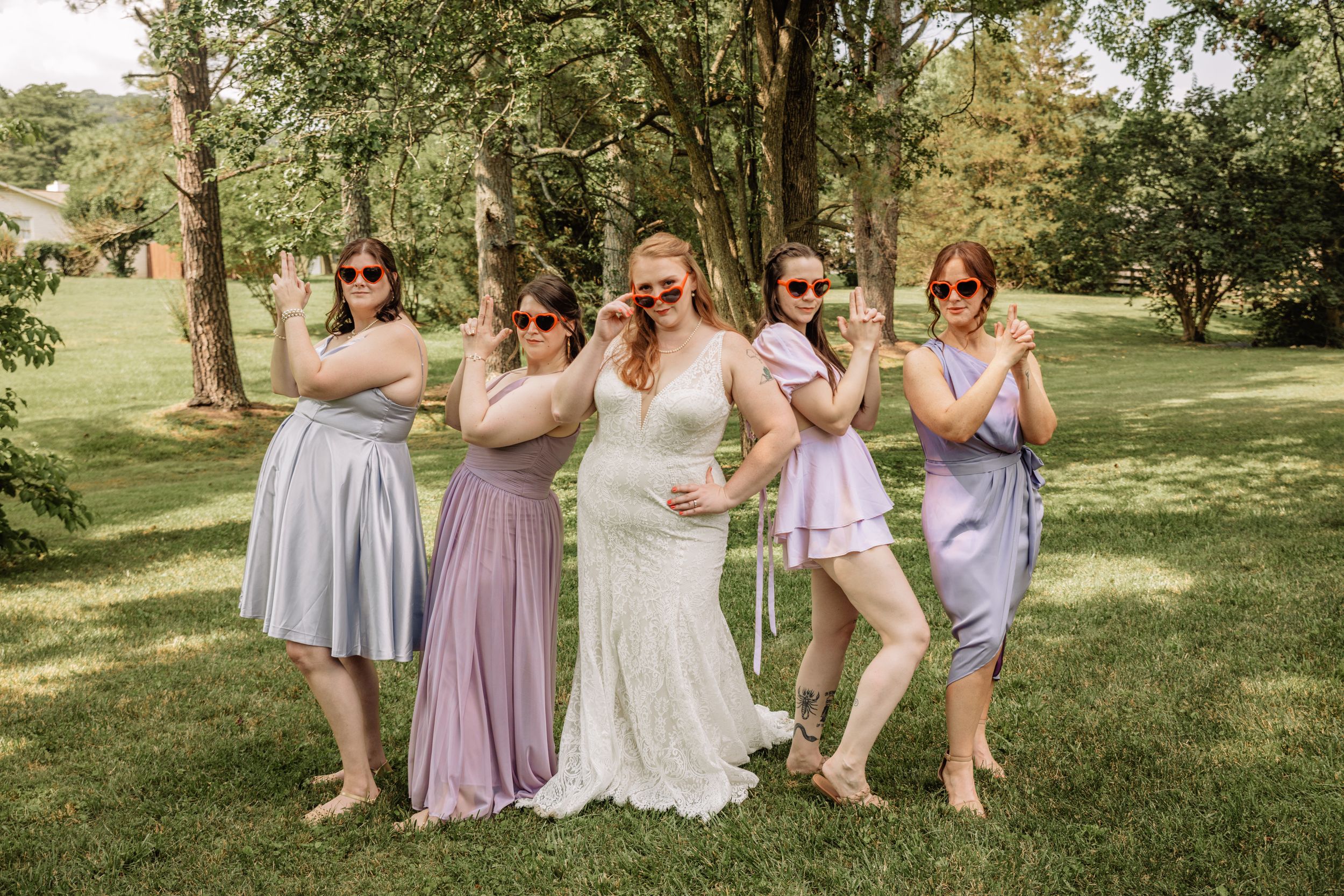 Wedding party members in coordinated purple and blue dresses pose on sunny lawn.