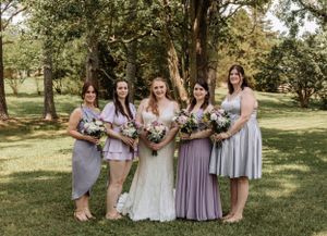 Wedding party in purple and grey dresses poses together on grass under trees.