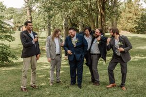 Wedding party members in suits gather playfully on grass under trees during an outdoor photo session.