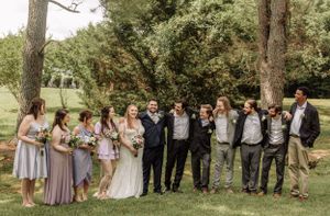 Large wedding party gathers for formal photo beneath pine trees on sunny afternoon.