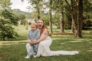 Bride in white gown kneels beside small child on grassy lawn with trees in background.