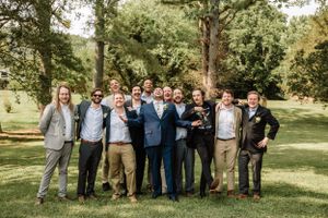 Wedding party members stand together outdoors on a grassy lawn with trees creating a natural backdrop.