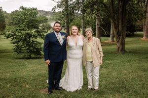 Wedding group gathers for formal photos under tall trees in a sunny outdoor setting.