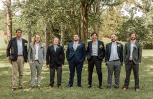 Wedding party groomsmen lined up outdoors in mixed formal attire against a wooded backdrop.