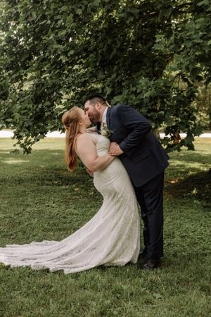 Wedding couple embraces in dramatic dip pose on grassy lawn with bride's lace dress trailing behind.