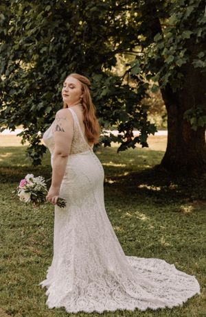 A lace wedding dress with a flowing train is displayed outdoors in a garden setting with natural lighting.